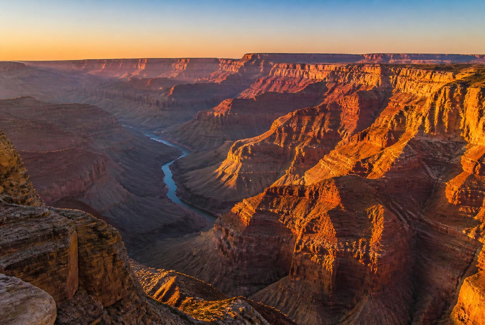 Layered red and ochre canyon walls at golden hour with the Colorado River winding through the gorge far below